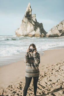 Junge Frau beim Fotografieren, während sie an einer Felsformation am Strand steht, Praia da Ursa, Lisboa, Portugal - FVSF00346