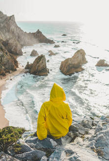 Rear view of woman wearing raincoat while sitting on rock looking at beach, Praia da Ursa, Lisboa, Portugal - FVSF00354