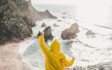 Young woman wearing raincoat with arms raised at Praia da Ursa, Lisboa, Portugal - FVSF00355