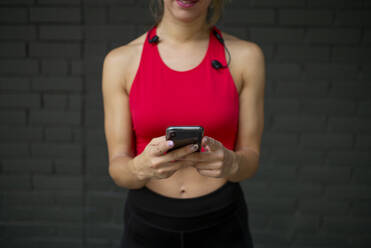 Weibliche Sportlerin in Sportkleidung, die ein Smartphone benutzt, während sie an der Wand steht - OCMF01247