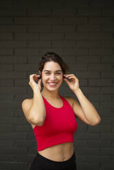 Lächelnde Sportlerin, die Musik über drahtlose Kopfhörer hört, während sie an einer Mauer steht - OCMF01248