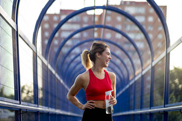Fröhliche Sportlerin, die eine Wasserflasche hält und wegschaut, während sie auf einer überdachten Brücke in der Stadt steht - OCMF01249