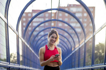 Lächelnde, selbstbewusste Sportlerin, die ein Smartphone benutzt, während sie auf einer überdachten Brücke in der Stadt steht - OCMF01250