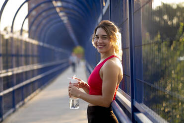 Lächelnde Sportlerin, die eine Wasserflasche hält, während sie auf einer überdachten Brücke in der Stadt steht - OCMF01251