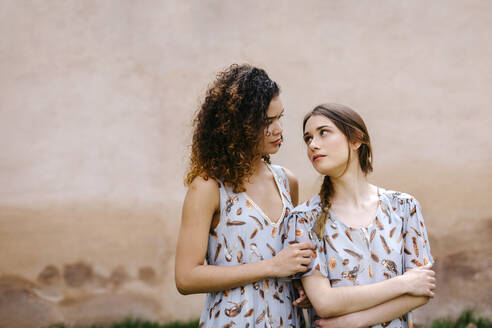 Loving sisters wearing dresses looking at each other while standing against old wall - TCEF00671