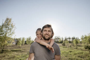 Smiling father piggybacking daughter while standing against clear sky in park during sunny day - EYAF01060