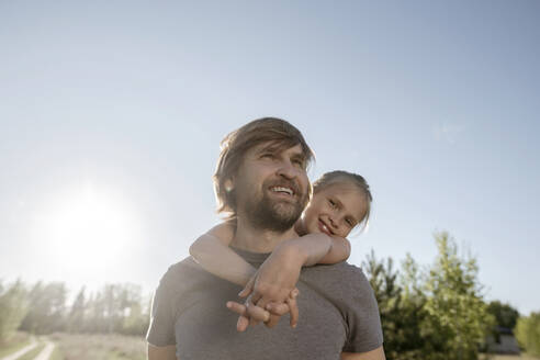 Glücklicher Vater, der seine Tochter im Huckepack nimmt, während er an einem sonnigen Tag im Park vor dem klaren Himmel steht - EYAF01061