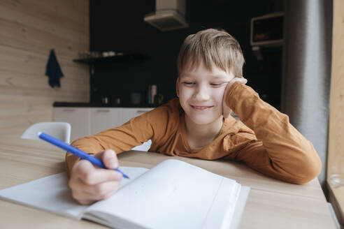 Smiling boy sitting at table writing into booklet at home - EYAF01068