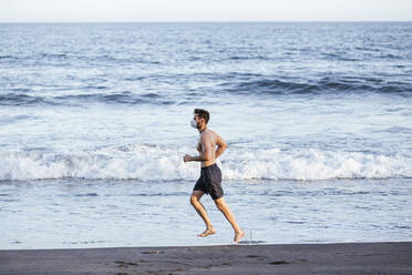 Shirtless mid adult man wearing face mask while running at beach during sunset - LJF01518