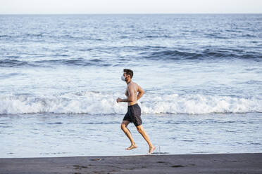 Shirtless Mitte erwachsenen Mann trägt Gesichtsmaske beim Laufen am Strand bei Sonnenuntergang - LJF01518