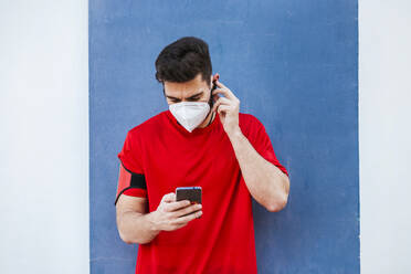 Mid adult man wearing mask using smart phone while standing against wall - LJF01526