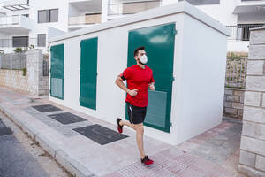 Mid adult man wearing face mask while running on footpath in city - LJF01527