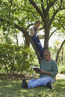 Entspannter Großvater und Enkelin im Garten, die ein Buch lesen und Erdbeeren essen - MCF00878