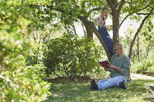 Entspannter Großvater und Enkelin im Garten, die ein Buch lesen und Erdbeeren essen - MCF00879