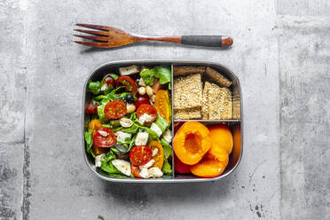 Lunchbox mit Rucolasalat mit bunten Tomaten, Mozzarella und Nüssen, Knäckebrot und Aprikosen, Holzgabel auf Betonfläche - SARF04587