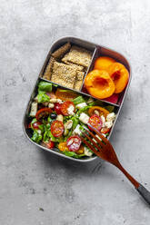 Lunchbox mit Rucolasalat mit bunten Tomaten, Mozzarella und Nüssen, Knäckebrot und Aprikosen, Holzgabel auf Betonfläche - SARF04588
