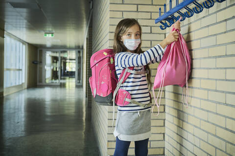 Mädchen mit Maske in der Schule hängt Tasche auf, lizenzfreies Stockfoto