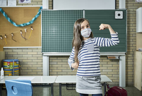 Portrait of girl wearing mask in classroom flexing muscles stock photo