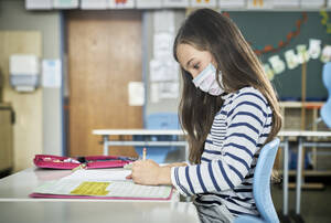 Mädchen mit Maske im Klassenzimmer schreibt in Arbeitsheft - DIKF00516