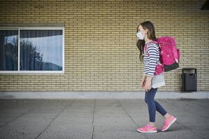 Girl wearing mask and schoolbag walking along building - DIKF00521