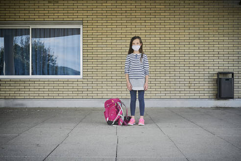 Girl with schoolbag wearing mask standing in front of building - DIKF00522