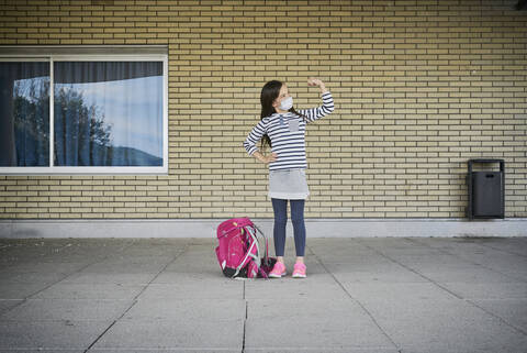 Mädchen mit Schultasche und Maske steht vor einem Gebäude und lässt ihre Muskeln spielen, lizenzfreies Stockfoto