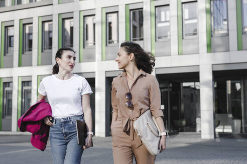 Multiethnische Geschäftsfrauen unterhalten sich im Gehen vor einem Bürogebäude - KMKF01395