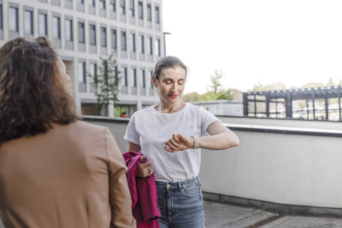Geschäftsfrau prüft Smartwatch, während sie mit einem Kollegen vor einem Bürogebäude steht - KMKF01401