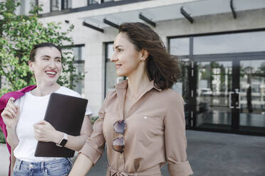 Fröhliche multiethnische Geschäftsfrauen, die sich unterhalten, während sie gegen ein Bürogebäude laufen - KMKF01403