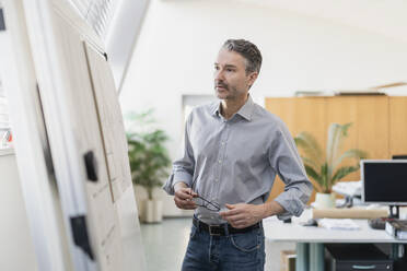 Gutaussehender reifer Geschäftsmann, der eine Brille hält, während er eine Strategie auf einer Tafel im Büro liest - DIGF11267