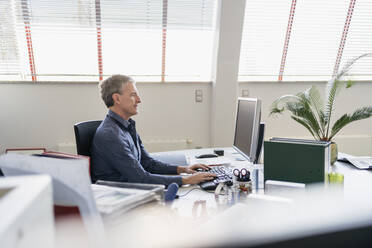 Side view of confident businessman using computer while sitting at desk in office - DIGF11270