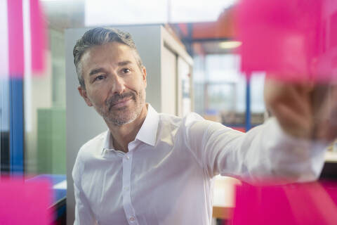 Selbstbewusster, gut aussehender männlicher Unternehmer, der eine Strategie plant, während er auf Haftnotizen auf einem Glas im Büro schaut, lizenzfreies Stockfoto