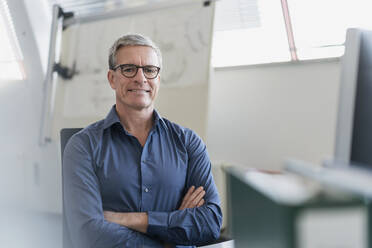 Porträt eines lächelnden reifen Geschäftsmannes, der mit verschränkten Armen am Schreibtisch im Büro sitzt - DIGF11300