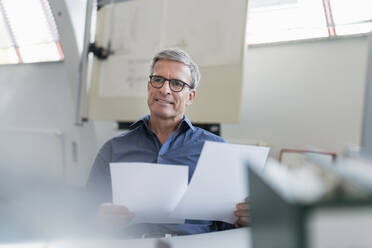 Lächelnder reifer männlicher Unternehmer, der am Schreibtisch im Büro sitzt und über ein Dokument nachdenkt - DIGF11305
