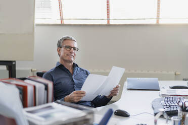 Nachdenklicher reifer Geschäftsmann schaut weg, während er mit Papieren am Schreibtisch im Büro sitzt - DIGF11306
