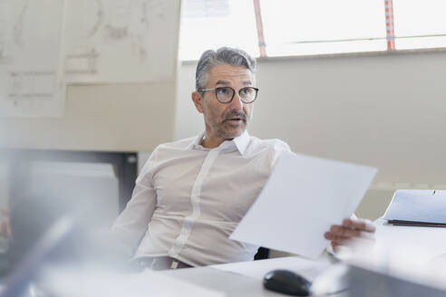 Confident businessman looking away with raised eyebrows while sitting with document at desk in office - DIGF11312