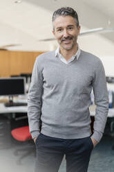 Portrait of smiling handsome businessman standing with hands in pockets at office - DIGF11317