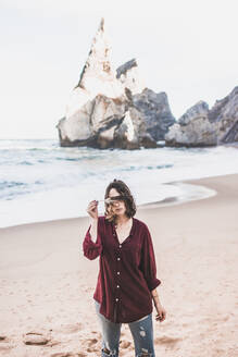 Junge Frau, die ihre Augen mit einer Feder bedeckt, während sie am Strand von Ursa steht, Region Lisboa, Portugal - FVSF00368