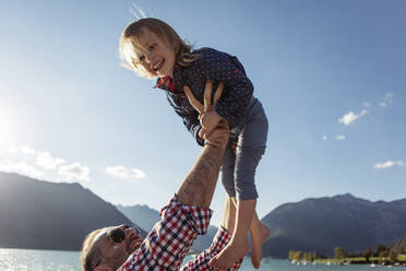 Father holding aloft cheerful daughter against blue sky at Achensee, Tyrol State, Austria - JLOF00398