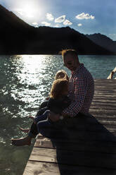 Father sitting with children on boardwalk by lake at Achensee, Tyrol State, Austria - JLOF00401