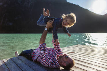 Man holding aloft cheerful daughter while lying on boardwalk by lake at Achensee, Tyrol State, Austria - JLOF00406