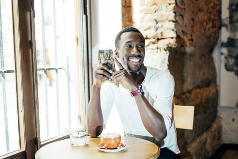 Lächelnder junger Mann mit Kaffee auf dem Tisch, der sein Smartphone hält und wegschaut, während er in einem Café sitzt - OCMF01253