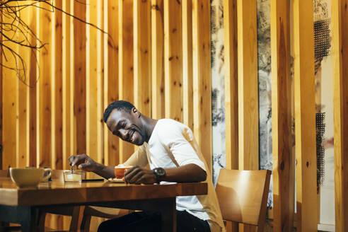 Lächelnder junger Mann, der den Kaffee auf dem Tisch betrachtet, während er in einem Café an der Wand sitzt - OCMF01256