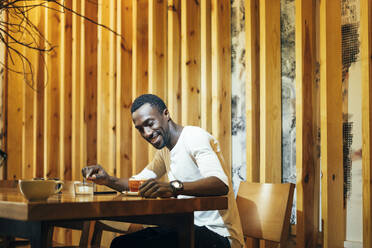 Lächelnder junger Mann, der den Kaffee auf dem Tisch betrachtet, während er in einem Café an der Wand sitzt - OCMF01257