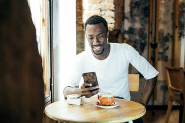Lächelnder junger Mann mit Kaffee auf dem Tisch, der sein Smartphone benutzt, während er in einem Café sitzt - OCMF01260