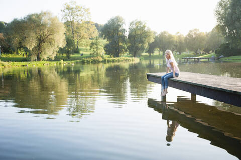 Young woman sitting on pier over lake in park during sunset stock photo