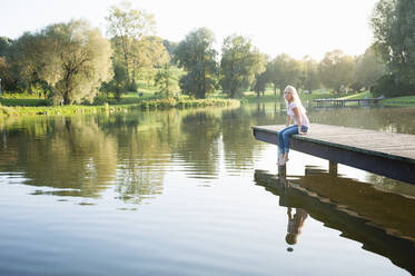 Junge Frau sitzt bei Sonnenuntergang auf einem Steg über einem See im Park - DIGF11385