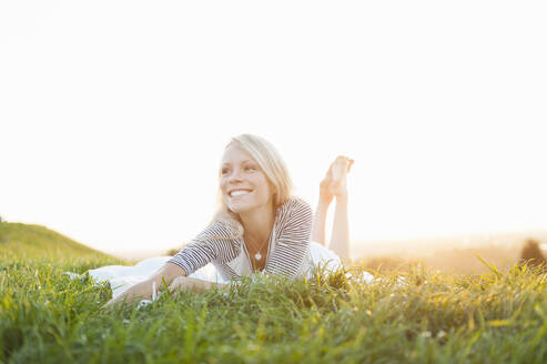 Glückliche, nachdenkliche junge Frau, die im Park bei Sonnenuntergang auf einer Wiese vor einem klaren Himmel liegt - DIGF11388
