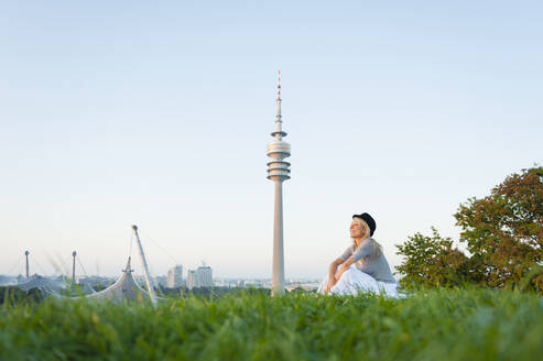 Nachdenkliche junge Frau sitzt auf einer Wiese mit dem Olympiaturm im Hintergrund in München, Deutschland - DIGF11389