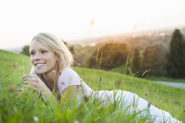 Lächelnde nachdenkliche junge Frau, die im Park bei Sonnenuntergang auf einer Wiese vor einem klaren Himmel liegt - DIGF11391